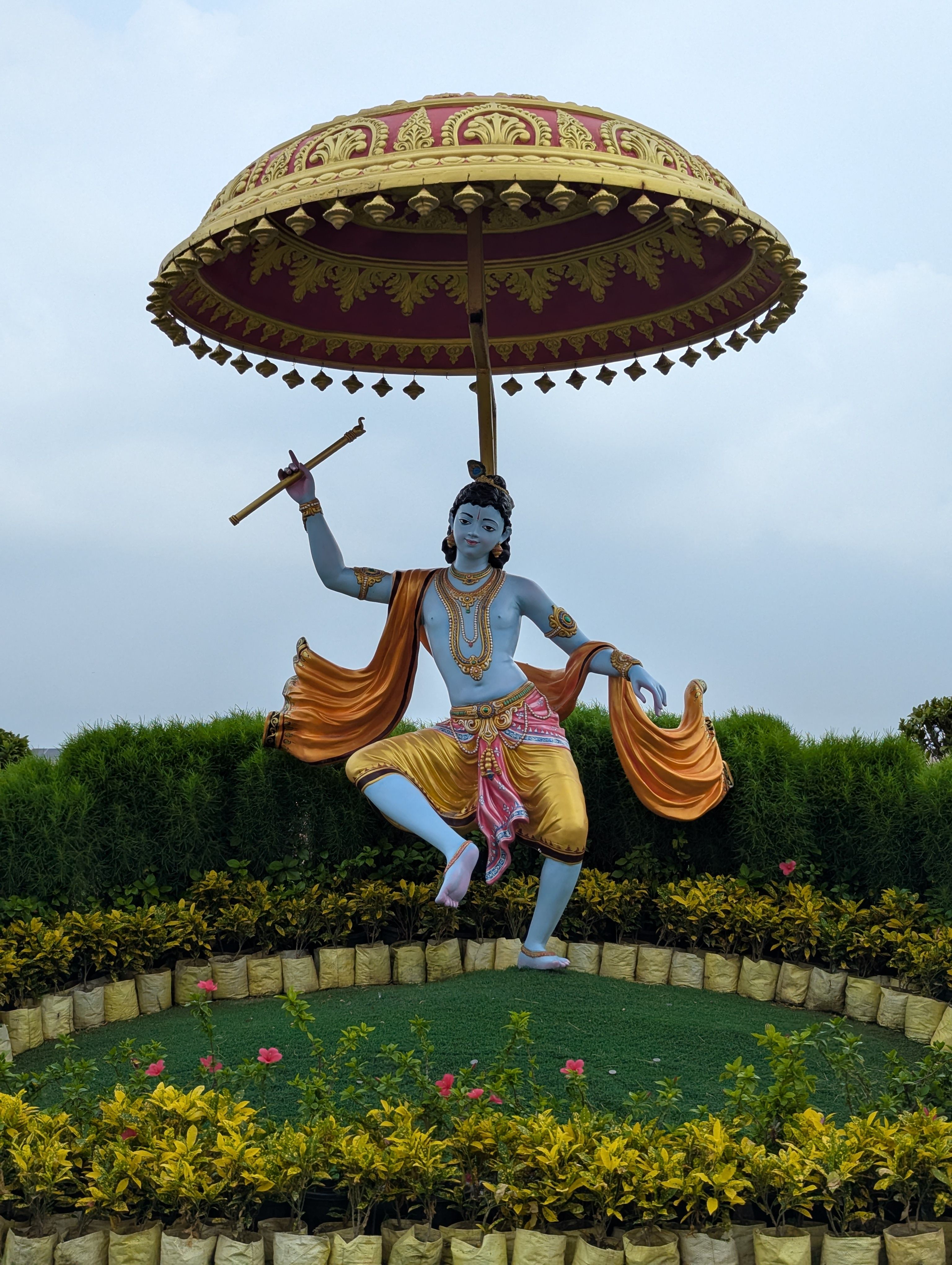 A vibrant statue of Lord Krishna with a flute under an ornate umbrella in a garden setting.