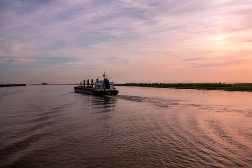 Free A serene sunset view of a cargo ship sailing on a peaceful river, highlighting tranquility and commerce. Stock Photo