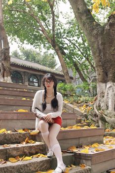 A young woman with glasses sits on stone steps surrounded by autumn leaves in a serene Nanjing garden.