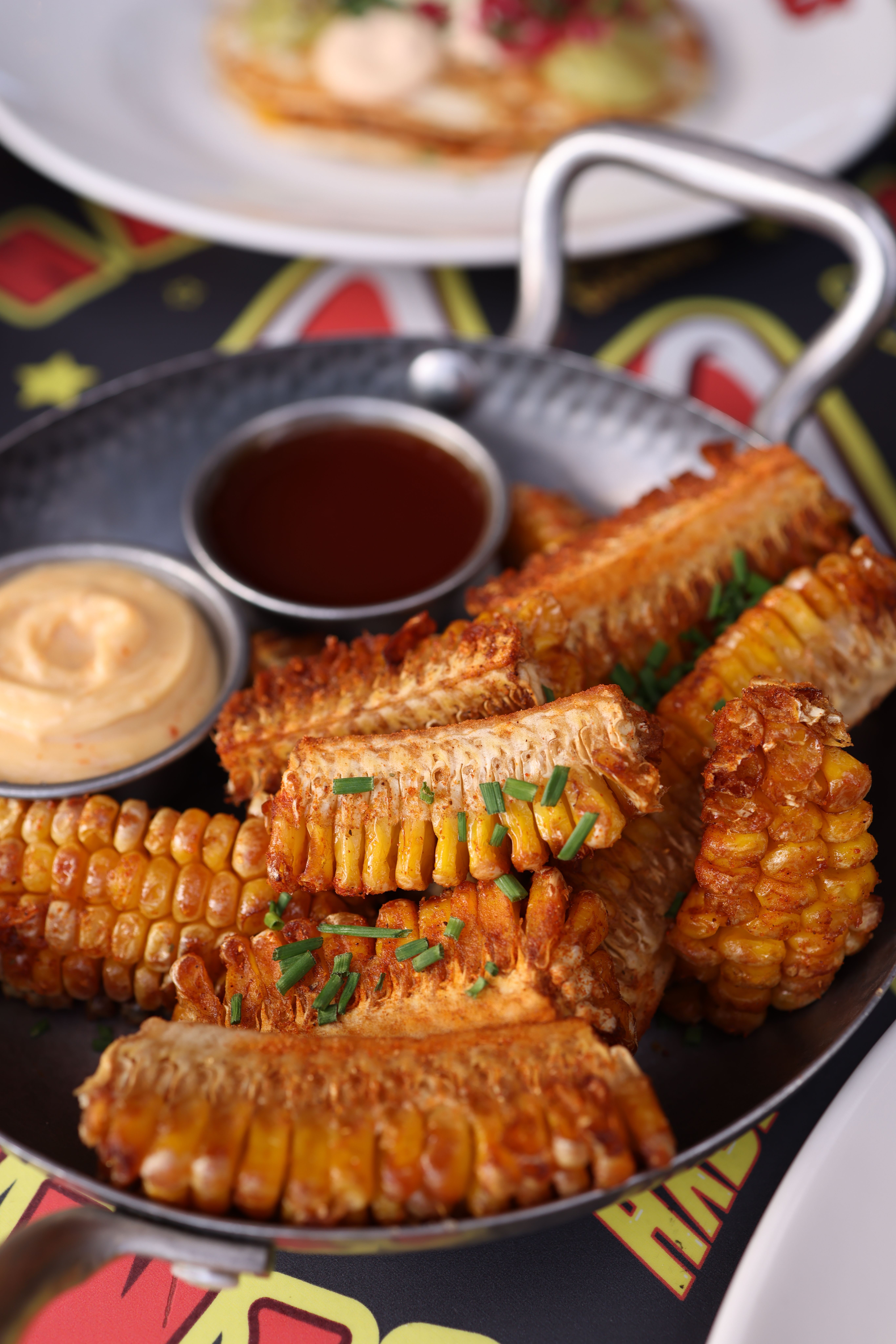 Charred corn ribs curled backward on a sheet pan, dusted with chili powder and cotija cheese, with a lime wedge and small bowl of chipotle crema alongside.