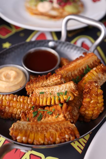 Charred corn ribs curled backward on a sheet pan, dusted with chili powder and cotija cheese, with a lime wedge and small bowl of chipotle crema alongside.