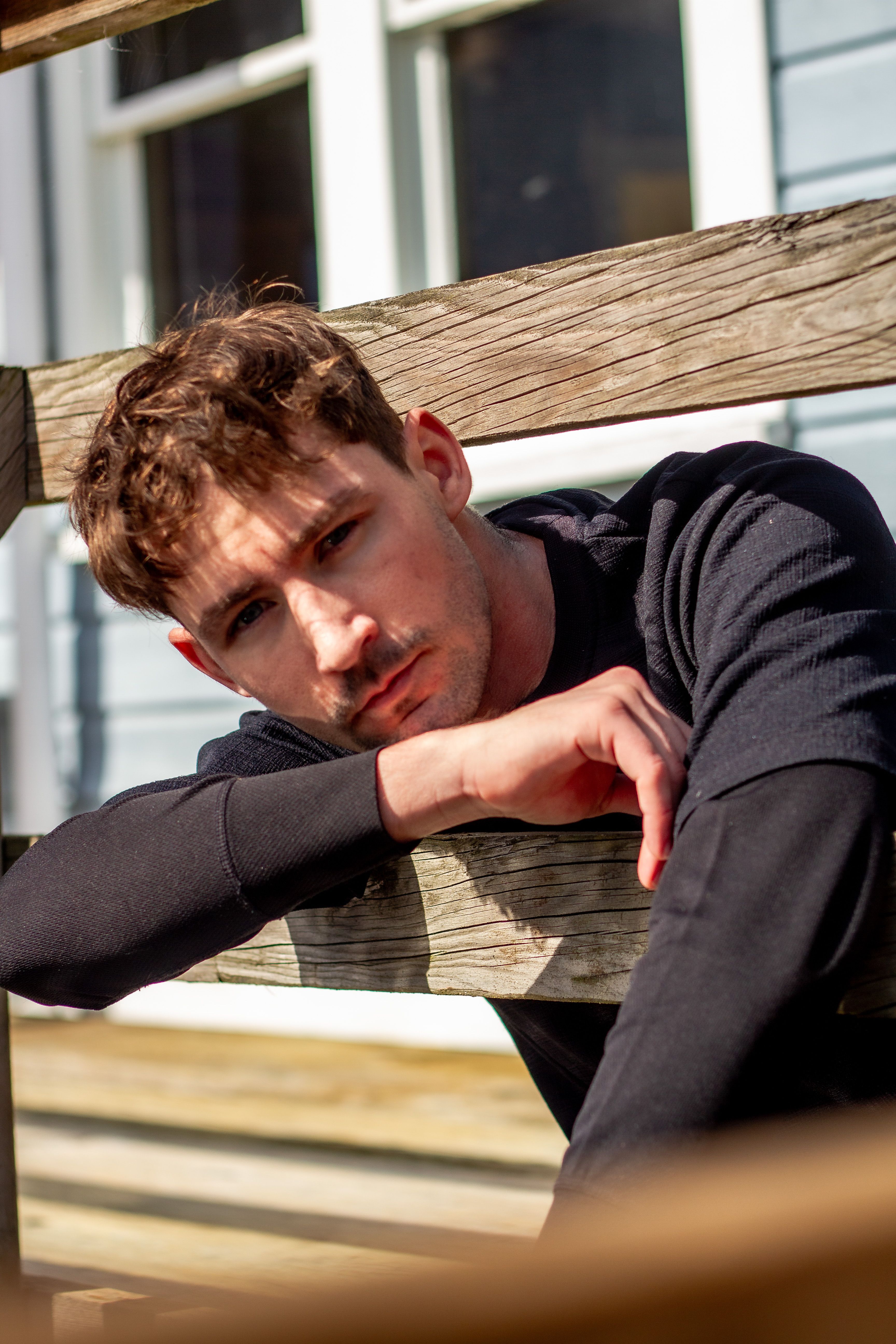 Young man in a relaxed pose outdoors, leaning on wooden railing, reflecting natural light and a warm atmosphere.