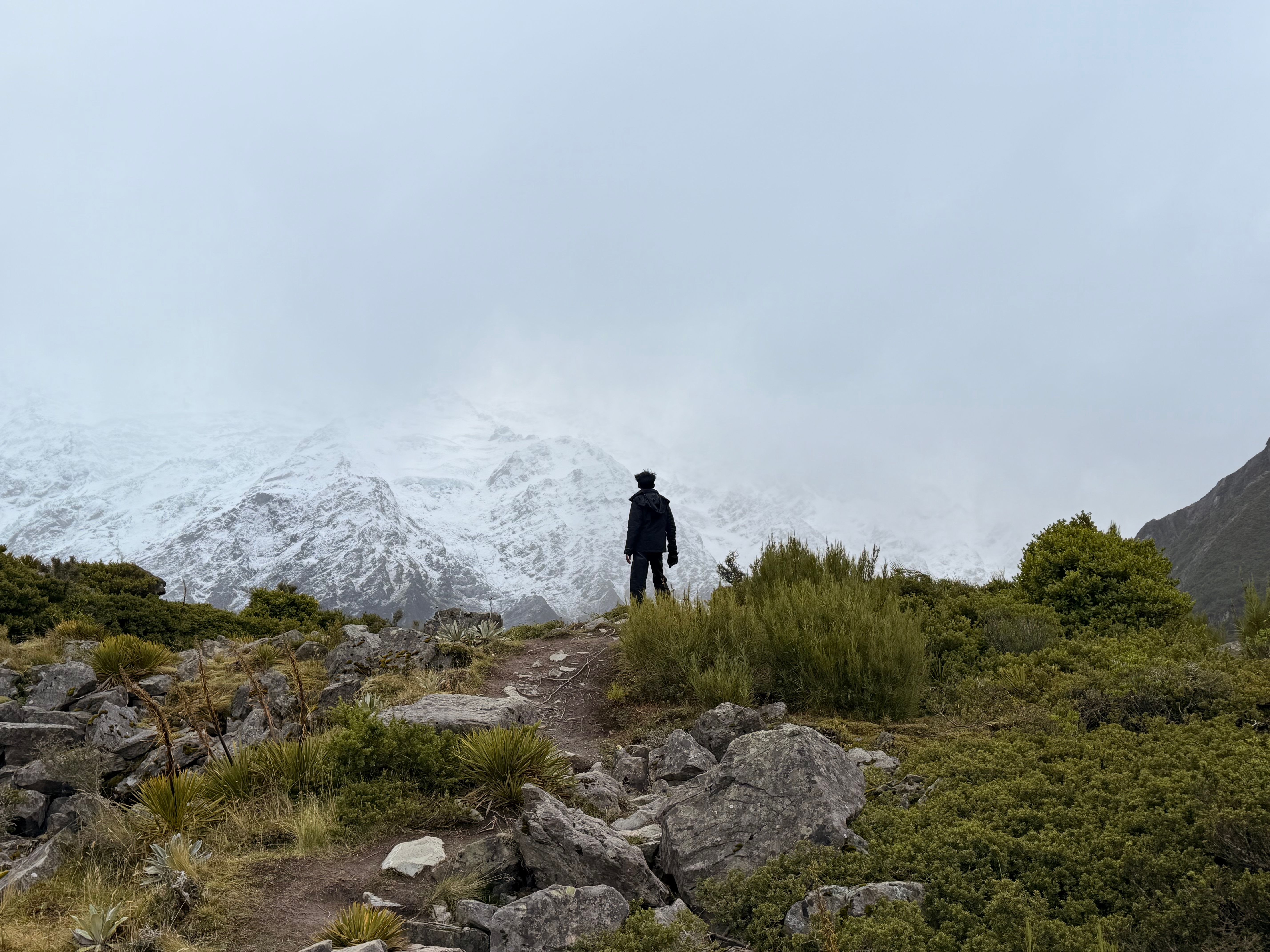 A lone trekker stands on a rocky trail overlooking snowy mountains in New Zealand's Canterbury Region.