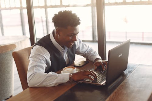 Positive young ethnic businessman working on laptop while sitting in cafe