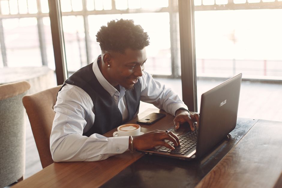 Side view of positive young African American male entrepreneur in formal clothes working on laptop while sitting at wooden table with cup of coffee in modern light cafe Side view of positive young African American male entrepreneur in formal clothes working on laptop while sitting at wooden table with cup of coffee in modern light cafe
