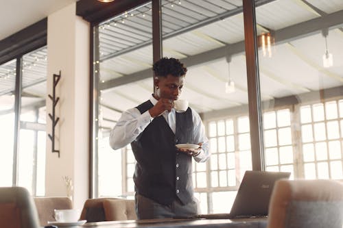 Serious young African American male entrepreneur in formal suit drinking cup of fresh coffee while working on laptop and standing near table in modern light cafe