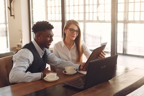 Smiling young multiethnic coworkers in formal clothes sitting with laptop and documents at wooden table while drinking coffee and discussing business ideas