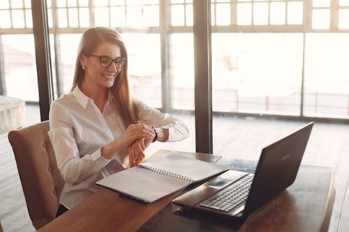 Positive young female entrepreneur in formal clothes and eyeglasses sitting at wooden table with laptop and copybook and checking time on wristwatch while working in light creative workspace