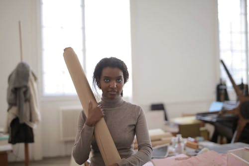 Young female ethnic worker in casual clothes standing near workbench with paper roll and looking at camera in modern workshop