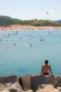 A crowded beach scene with swimmers and a scenic backdrop of hills under a clear summer sky.