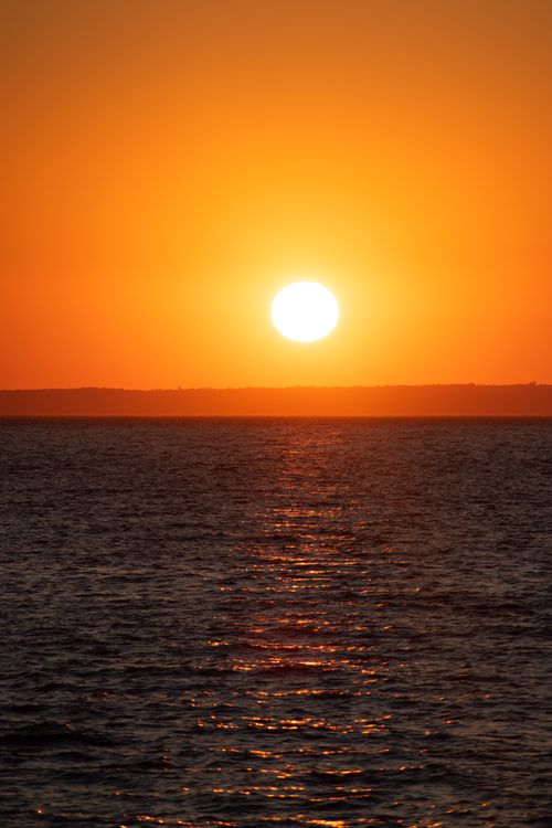 Free A vibrant orange sunset casts reflections over a calm sea, creating a serene atmosphere. Stock Photo