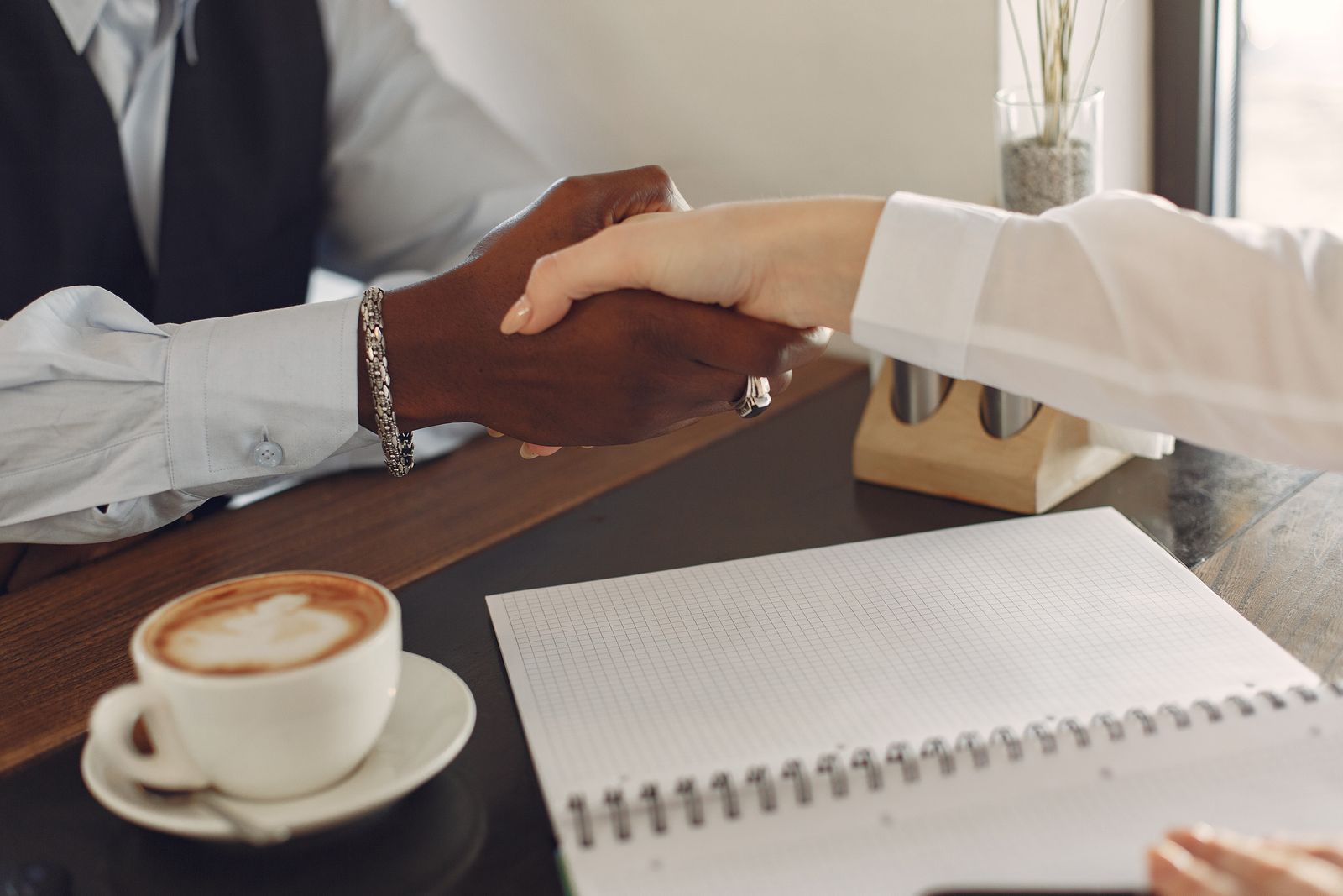 Two people shaking hands across a table beside a cup of coffee and an open notebook
