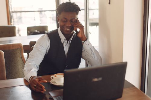 Free Smiling man in a relaxed office setting enjoying a phone conversation while working on a laptop with coffee. Stock Photo
