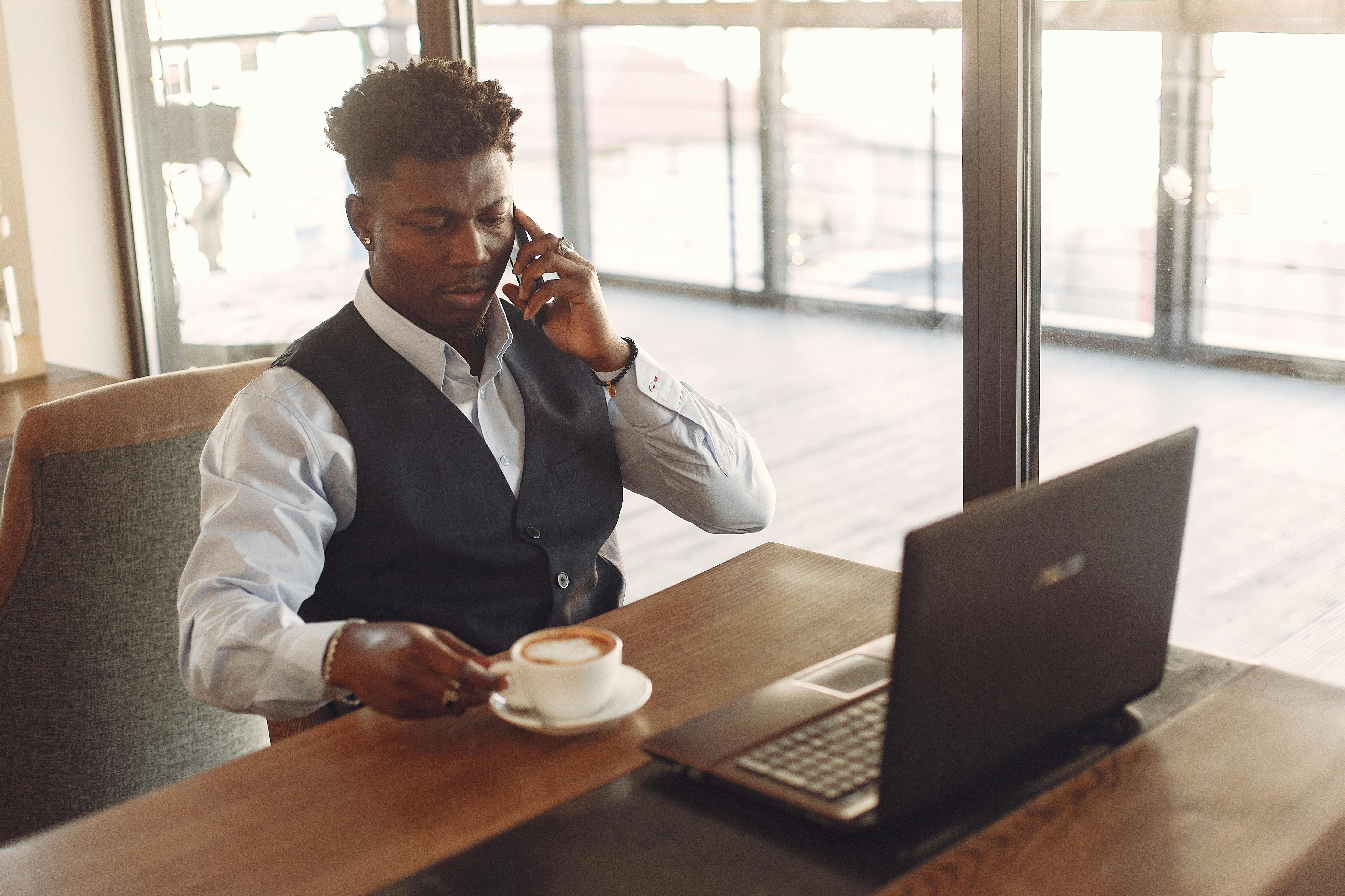 A businessman sits at a desk using a phone, drinking coffee, and working on a laptop.