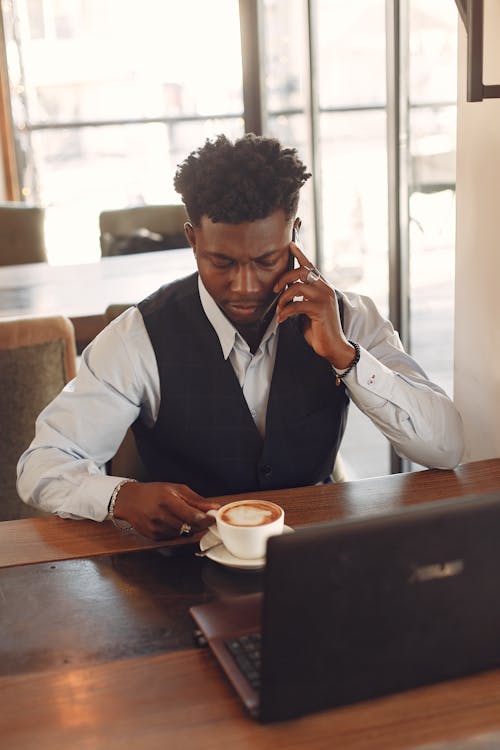 Top view of focused male entrepreneur in formal wear speaking on phone while drinking coffee in front of netbook in creative cafeteria