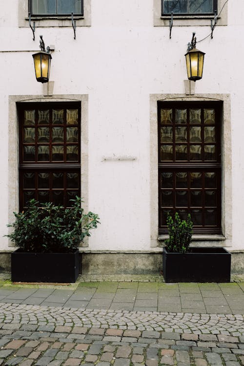 Exterior of aged stone house with potted plants and lighted lanterns above ornamental doorways on cobblestone street