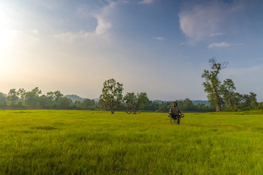A serene landscape with a man sitting in a vast, vibrant green field under a clear sky.