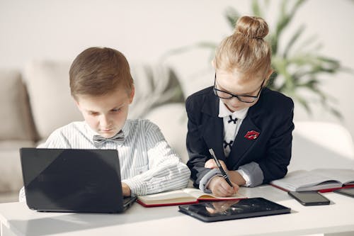 Cute busy children working on laptop and writing in notebook in light workspace