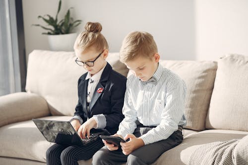 Concentrated children coworkers in formal clothing and glasses browsing tablet and laptop while working on project together in cozy  workspace