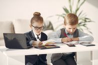 Cute business children in uniform using laptop and writing information in planners while making up startup strategy in modern workspace