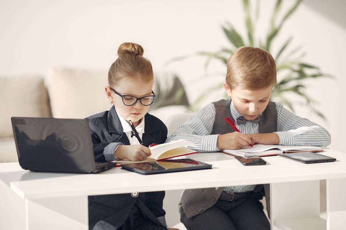 Free Cute business children in uniform using laptop and writing information in planners while making up startup strategy in modern workspace Stock Photo