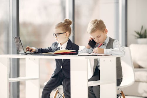 Little preschool manager in uniform talking on phone and taking notes in planner while girl colleague in formal wear and glasses using netbook in modern studio