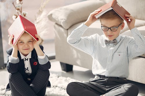 Smiling little students in formal clothes gathering on floor near sofa and playing games with textbooks while studying together at home