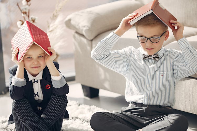 Positive Cute Kids Sitting With Books On Heads Near Sofa In Cozy Living Room