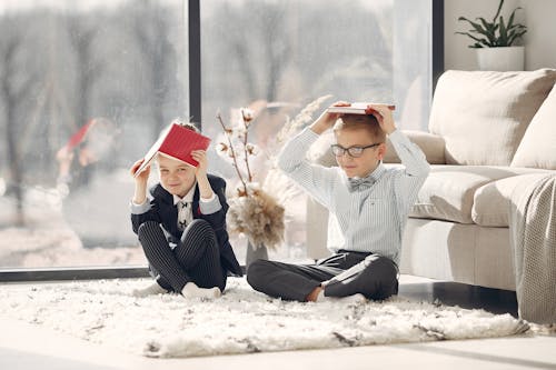 Full body happy little students in uniform and eyeglasses sitting on floor near sofa and window putting books on head while staying in modern room