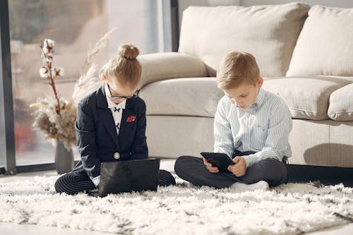 Serious kids in formal wear and eyeglasses sitting on carpet surfing table and laptop while working remotely in living room