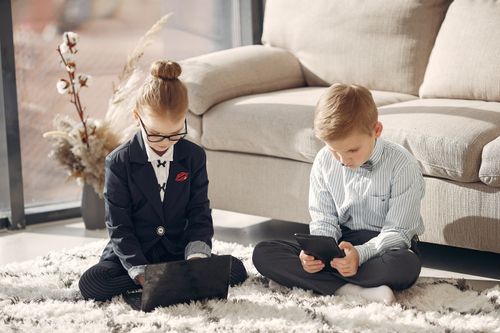 Free Full body of concentrated kids in uniform using devices while studying  online and sitting on carpet near sofa in cozy living room Stock Photo