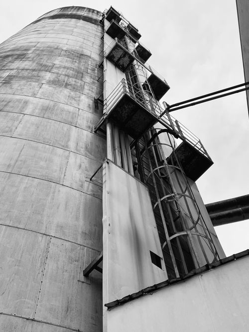 Free Black and white photo of a towering industrial silo with steel ladders and platforms, skyward view. Stock Photo