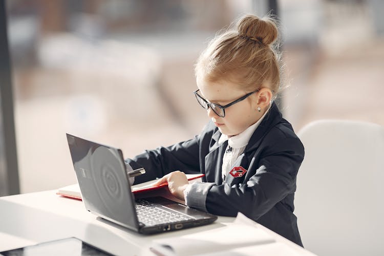 Side View Of Cute Little Businesswoman Taking Notes In Daily Planner Using Laptop In Office