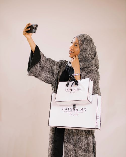 Free Young woman in abaya taking a selfie, holding Laihaa.NG shopping bags, indoors. Stock Photo