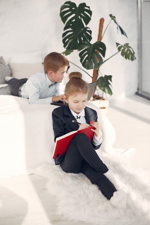 Cute boy wearing formal clothes using laptop while lying in bed together with adorable girl wearing formal suit sitting on floor and writing in notebook in bedroom with white interior at modern apartment