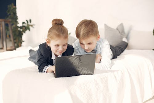 Little adorable boy and girl in formal clothing lying on bed and browsing netbook during rest in modern bedroom with white interior and smiling looking at screen
