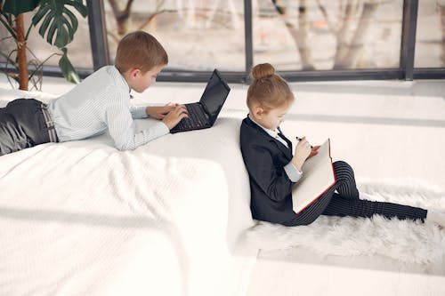 Free Side view of attentive cute boy using laptop while lying in bed together with adorable serious girl sitting on fluff carpet near bed and taking notes in diary in modern apartment with white interior Stock Photo
