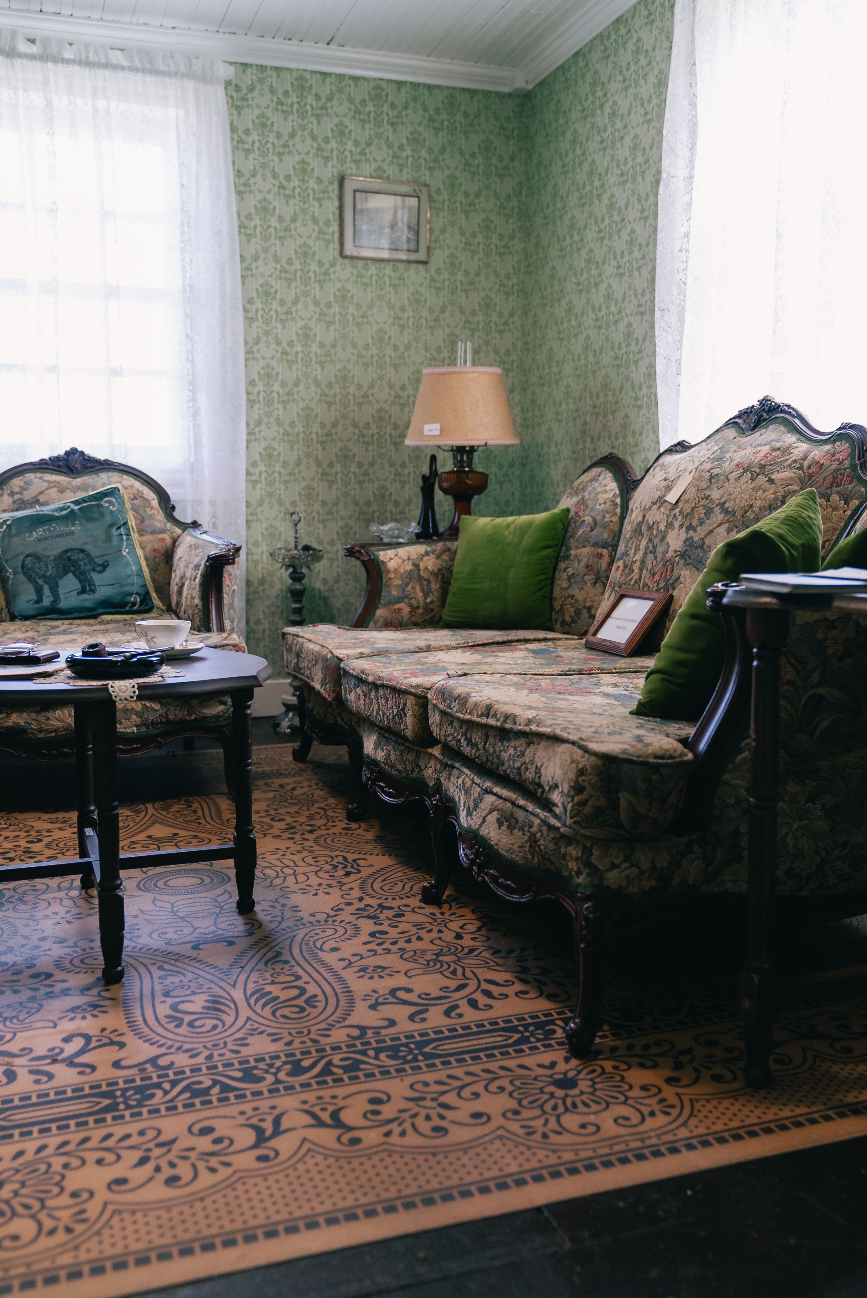 A vintage living room with a floral antique sofa, green patterned wallpaper, and a lamp on a side table.