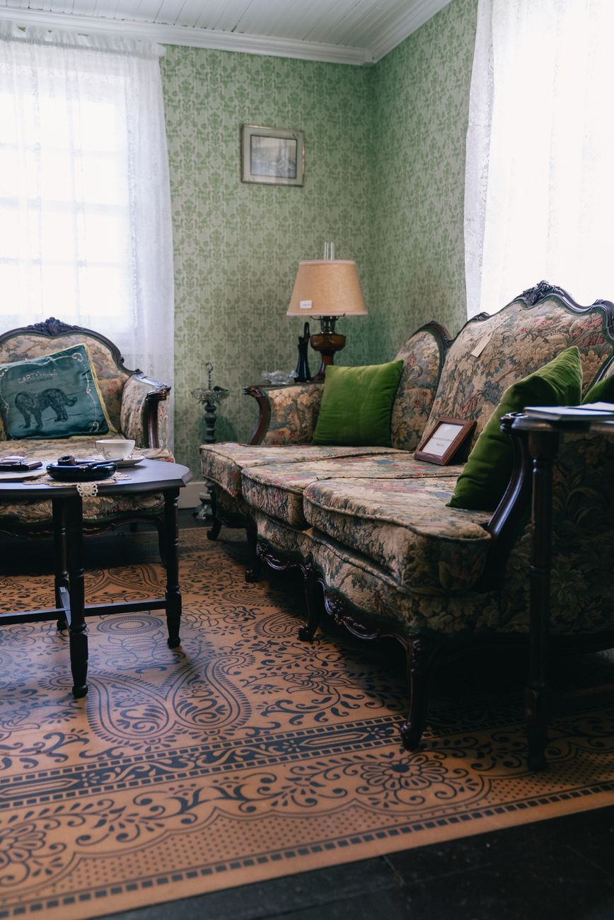 A vintage living room with a floral antique sofa, green patterned wallpaper, and a lamp on a side table.