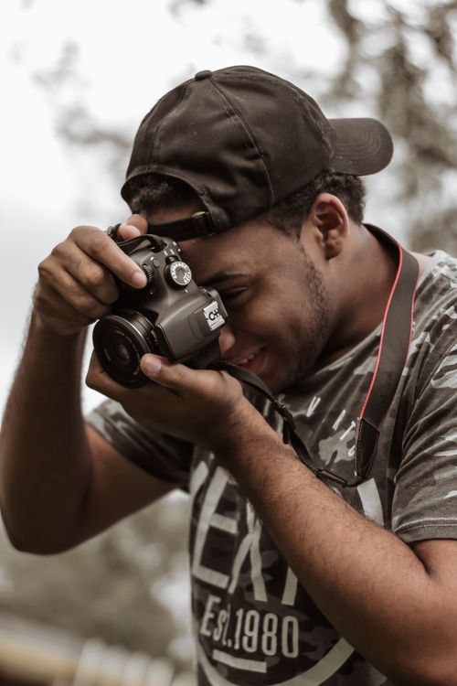 Free A young man wearing a cap captures a moment with his DSLR camera outdoors, smiling as he focuses on the shot. Stock Photo