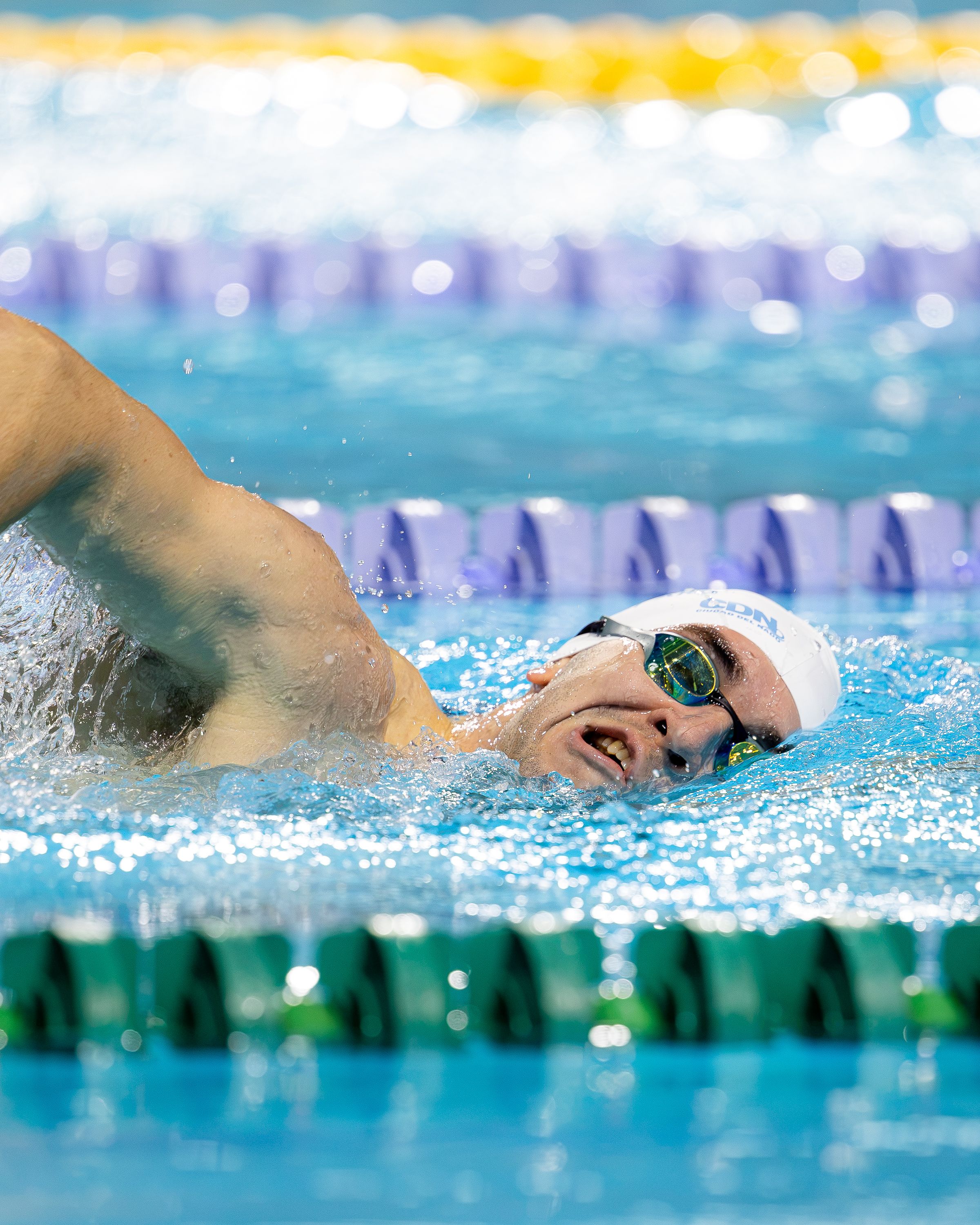 Male athlete swimming freestyle in competition pool indoors, wearing swim cap and goggles.
