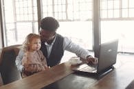 Cheerful young African American man looking after little girl in cafe using laptop