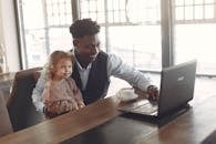 Cheerful black man in formal wear sitting together with cute child of diverse colleague at table with cup of coffee and using laptop in modern cafeteria during daytime