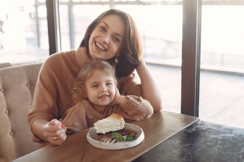 Free Young woman in casual outfit sitting at desk and eating piece of cake together with little adorable girl while spending time at cafeteria and smiling looking at camera Stock Photo