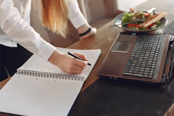 From above of crop anonymous female freelancer writing with pen in copybook while standing near table with laptop and plate of food for lunch in cafeteria