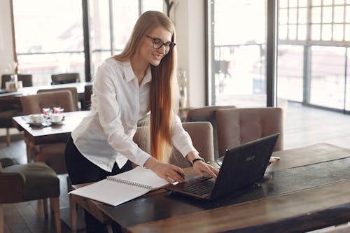 Free Happy female freelancer wearing white formal shirt and eyeglasses standing near table with laptop and copybook working on business project in modern cafeteria Stock Photo