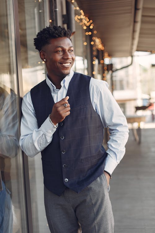 Smiling young handsome African American male standing on urban city street leaning on glass wall of cafeteria with hand in pocket and looking away