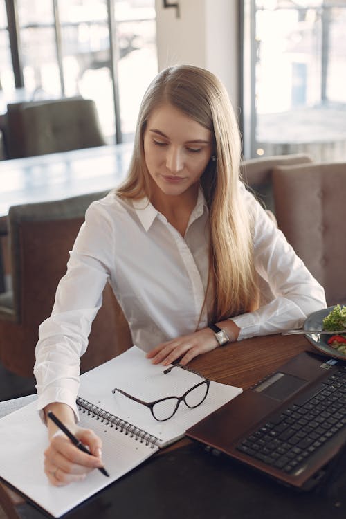 Young woman writing in notebook while working on laptop