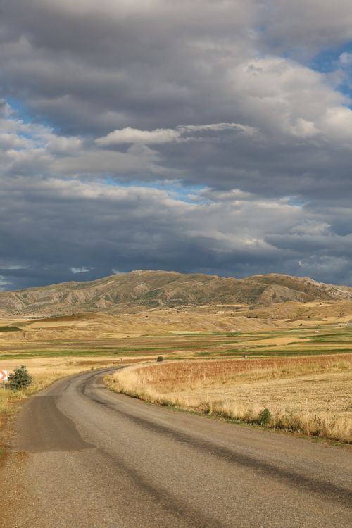 Free Curving road through a picturesque field with distant mountains under a dramatic sky. Stock Photo