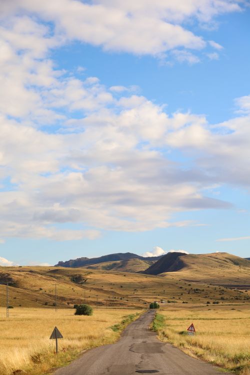 Free A picturesque rural road stretching through golden fields under a vibrant blue sky with clouds. Stock Photo